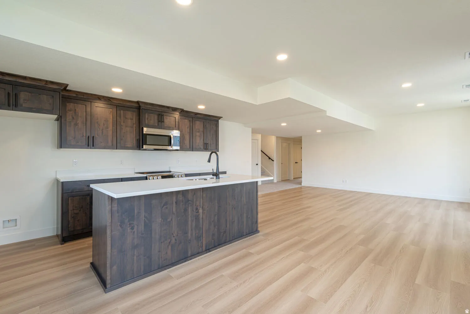 Kitchen with dark wood finish cabinets, stainless steel microwave, light wood-type flooring, a kitchen island with sink, and open floor plan