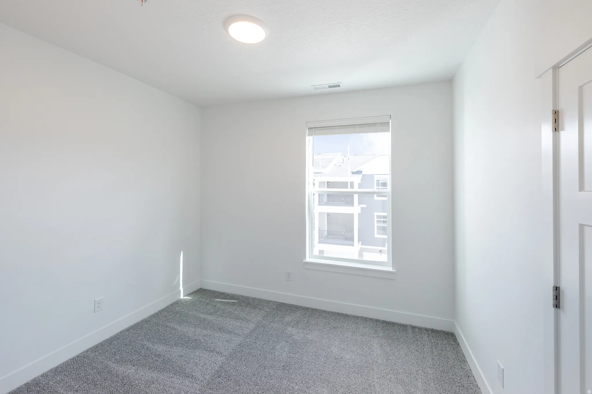 Recessed ceiling lighting, white walls, gray patterned carpet, single window with white trim, and a white paneled door