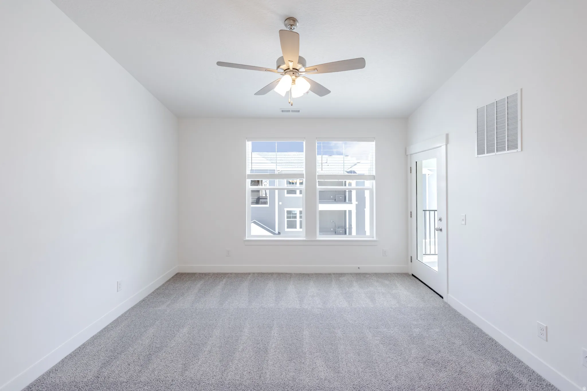 Neutral-toned carpeting, white walls, two large windows, a ceiling fan with integrated lighting, and a glass-paneled door