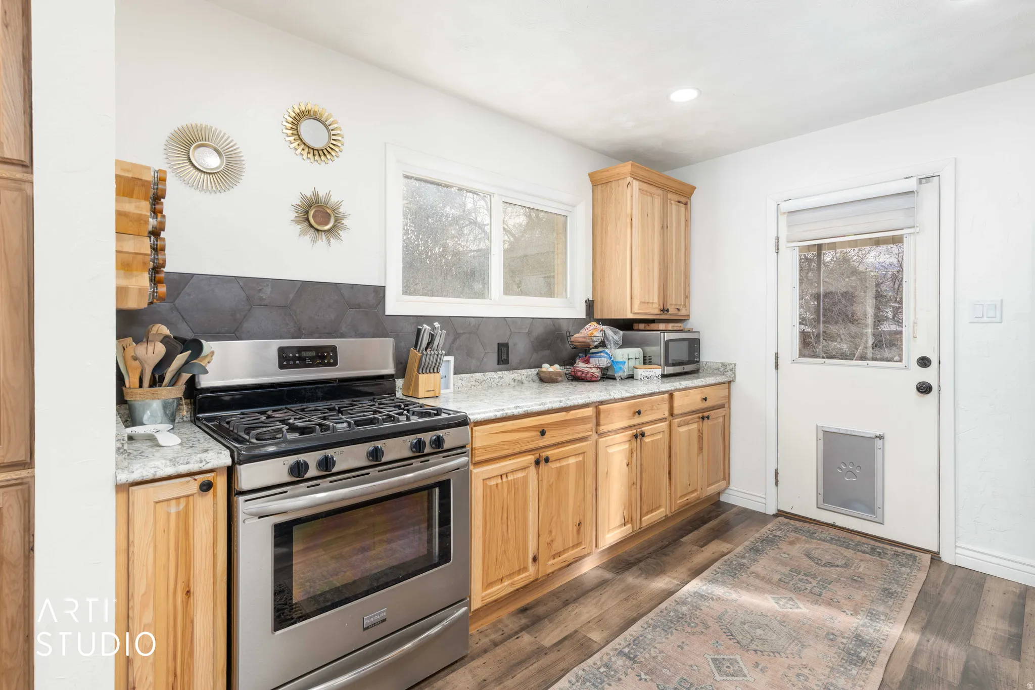 Kitchen featuring stainless steel appliances, decorative backsplash, light stone counters, light wood finish cabinetry, and dark wood-style flooring