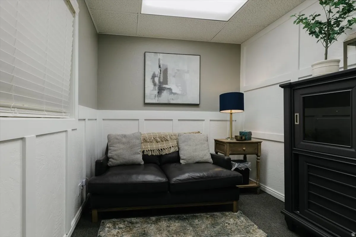 Living area with a decorative wall, a drop ceiling, dark carpet, and a wainscoted wall