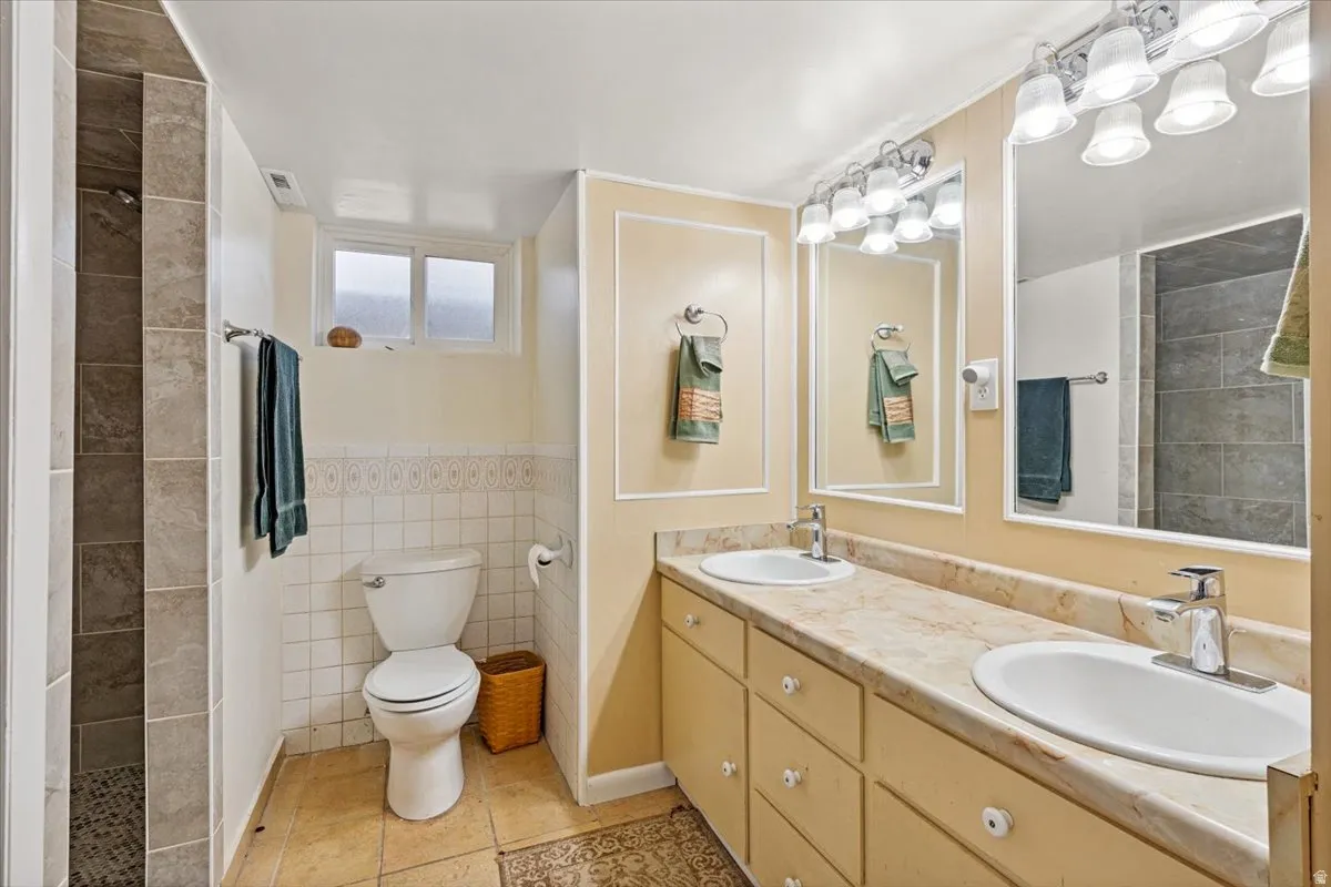 Bathroom featuring double vanity, tile walls, tiled shower, and a wainscoted wall