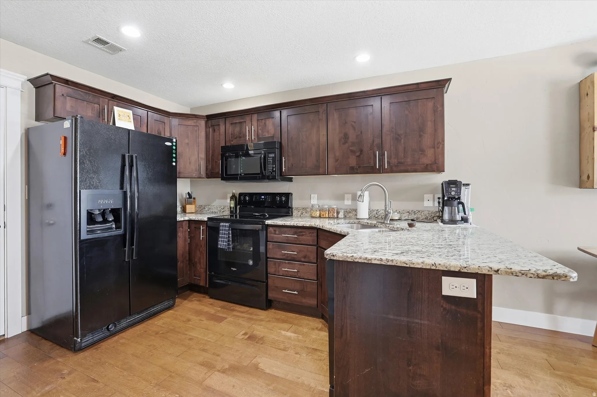 Kitchen featuring dark wood finish cabinets, a peninsula, black appliances, light stone countertops, and light wood-style floors