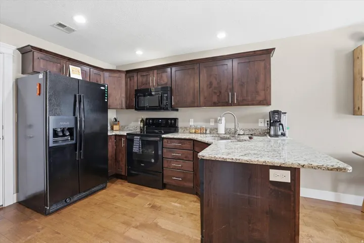 Kitchen featuring dark wood finish cabinets, a peninsula, black appliances, light stone countertops, and light wood-style floors