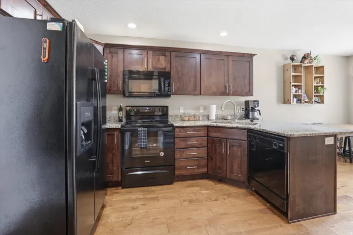Kitchen featuring black appliances, dark wood finish cabinets, a peninsula, light stone counters, and recessed lighting