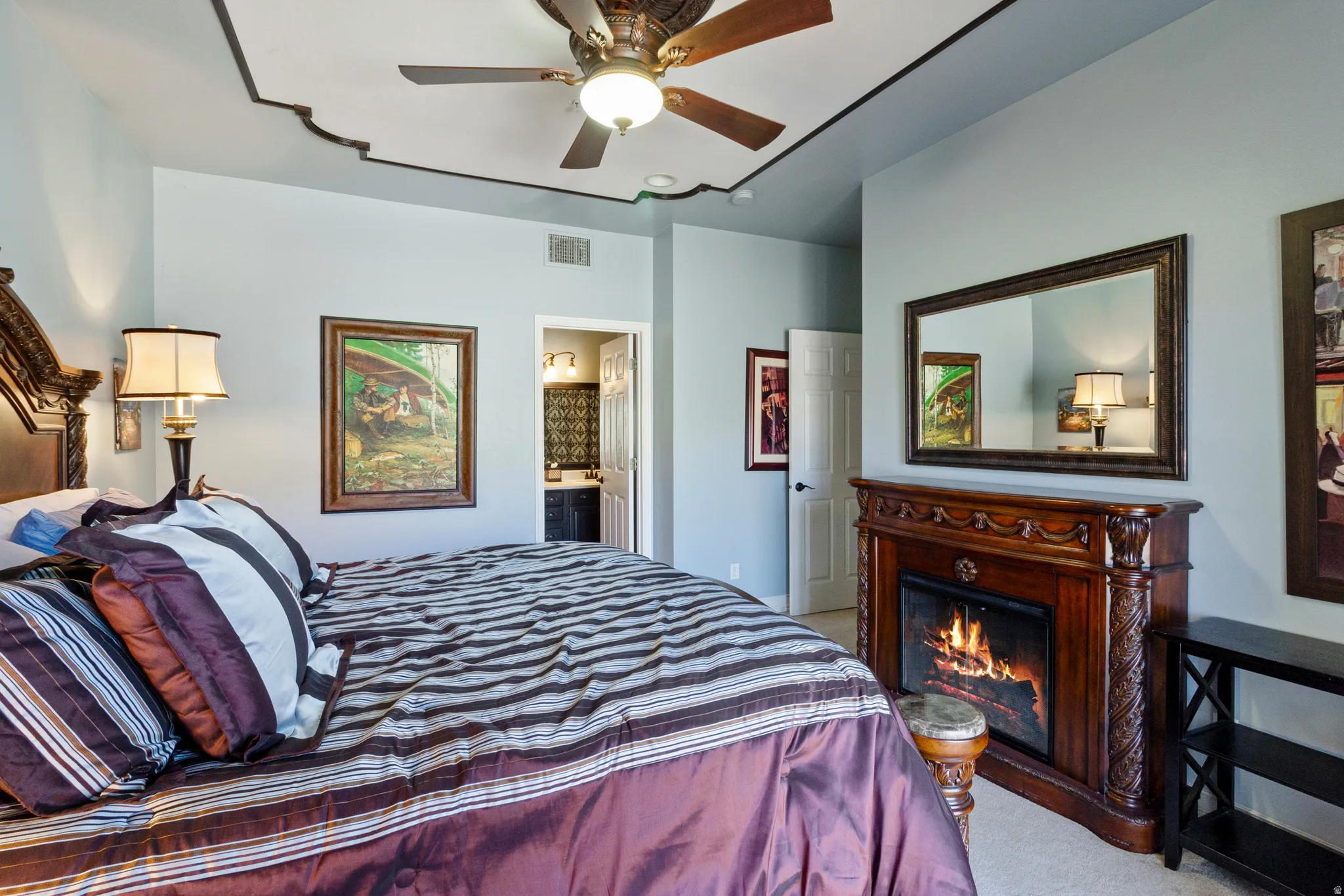 Bedroom featuring ceiling fan, light colored carpet, connected bathroom, and a lit fireplace