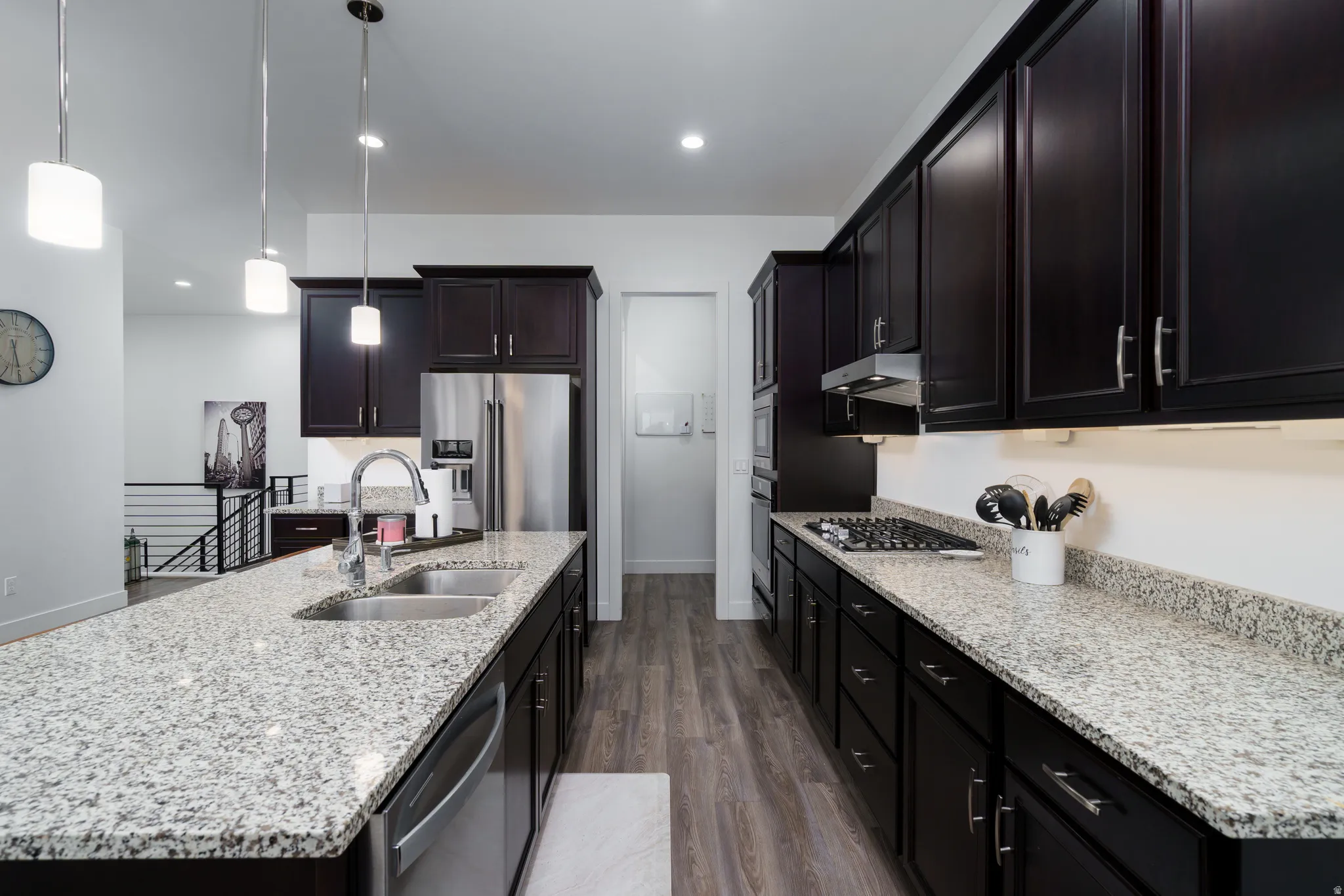 Kitchen featuring stainless steel appliances, light stone countertops, an island with sink, dark wood-style floors, and decorative light fixtures