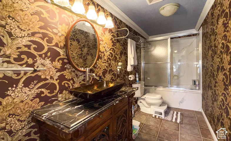 Full bathroom featuring wallpapered walls, bath / shower combo with glass door, ornamental molding, vanity, and dark tile patterned floors