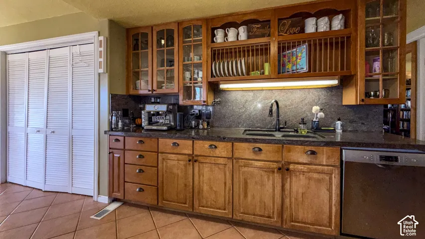 Kitchen with stainless steel dishwasher, wood finish cabinetry, tasteful backsplash, light tile patterned flooring, and glass insert cabinets