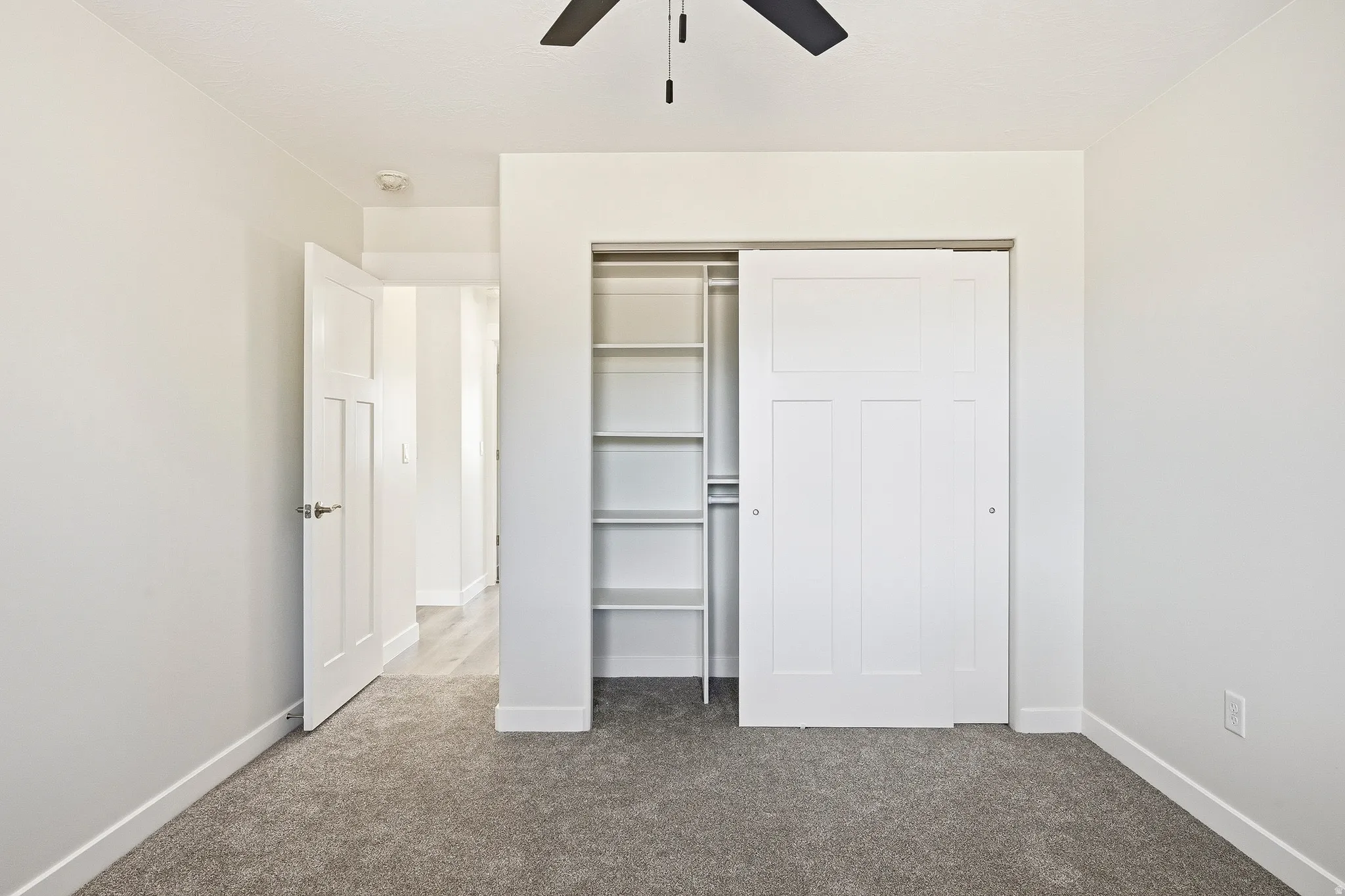 Plush carpet flooring, white painted walls, ceiling fan, sliding closet door with built-in shelving, and a paneled interior door