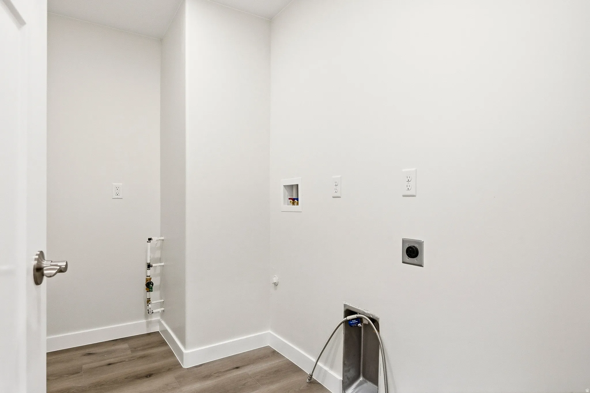 Wood-finish flooring with white baseboards, utility sink with flexible hose, appliance hookups, and a white interior door with brushed nickel hardware