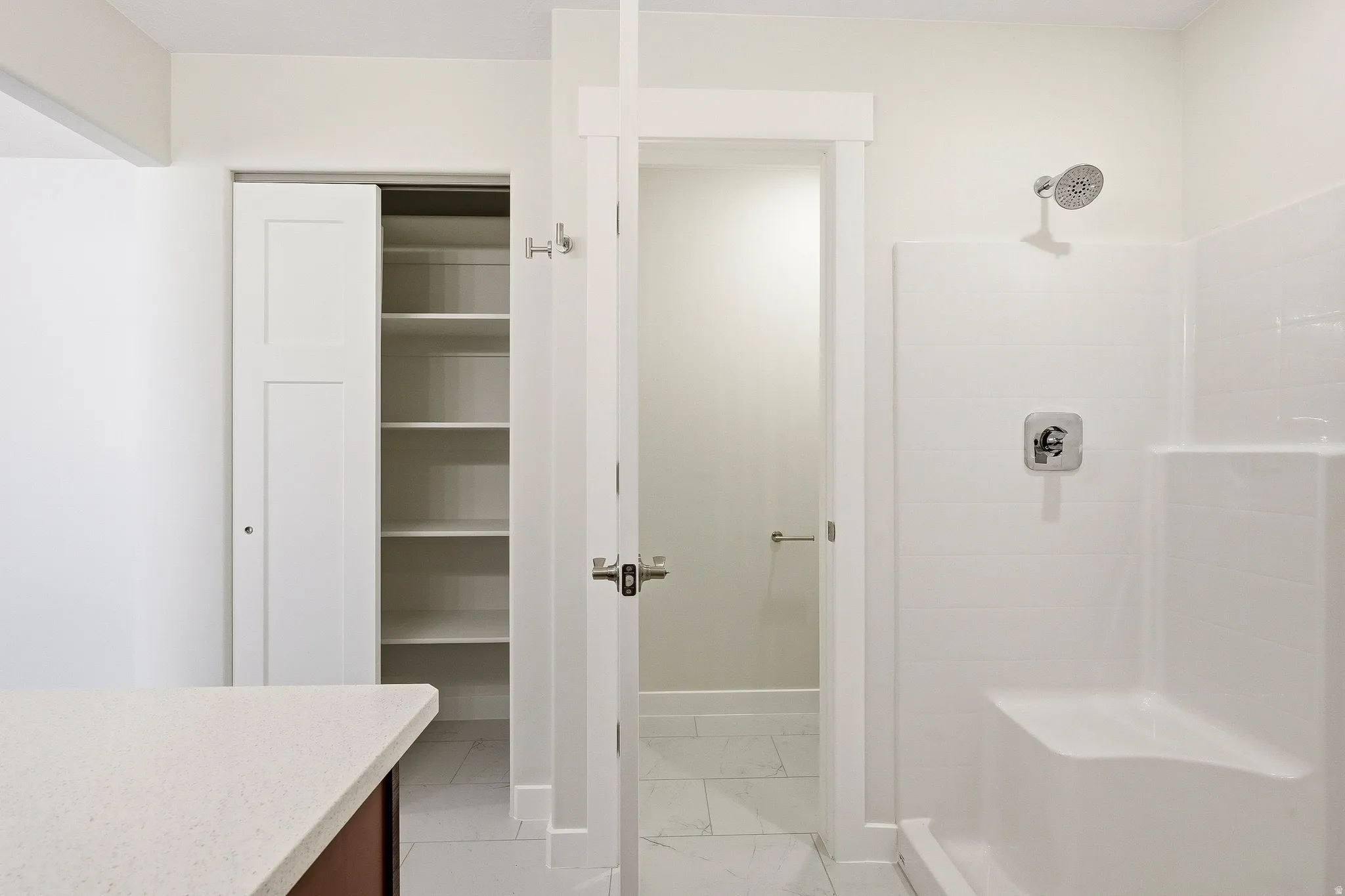 White solid surface countertop, built-in shelving, white sliding closet door, white shower/tub combination, and chrome shower fixtures