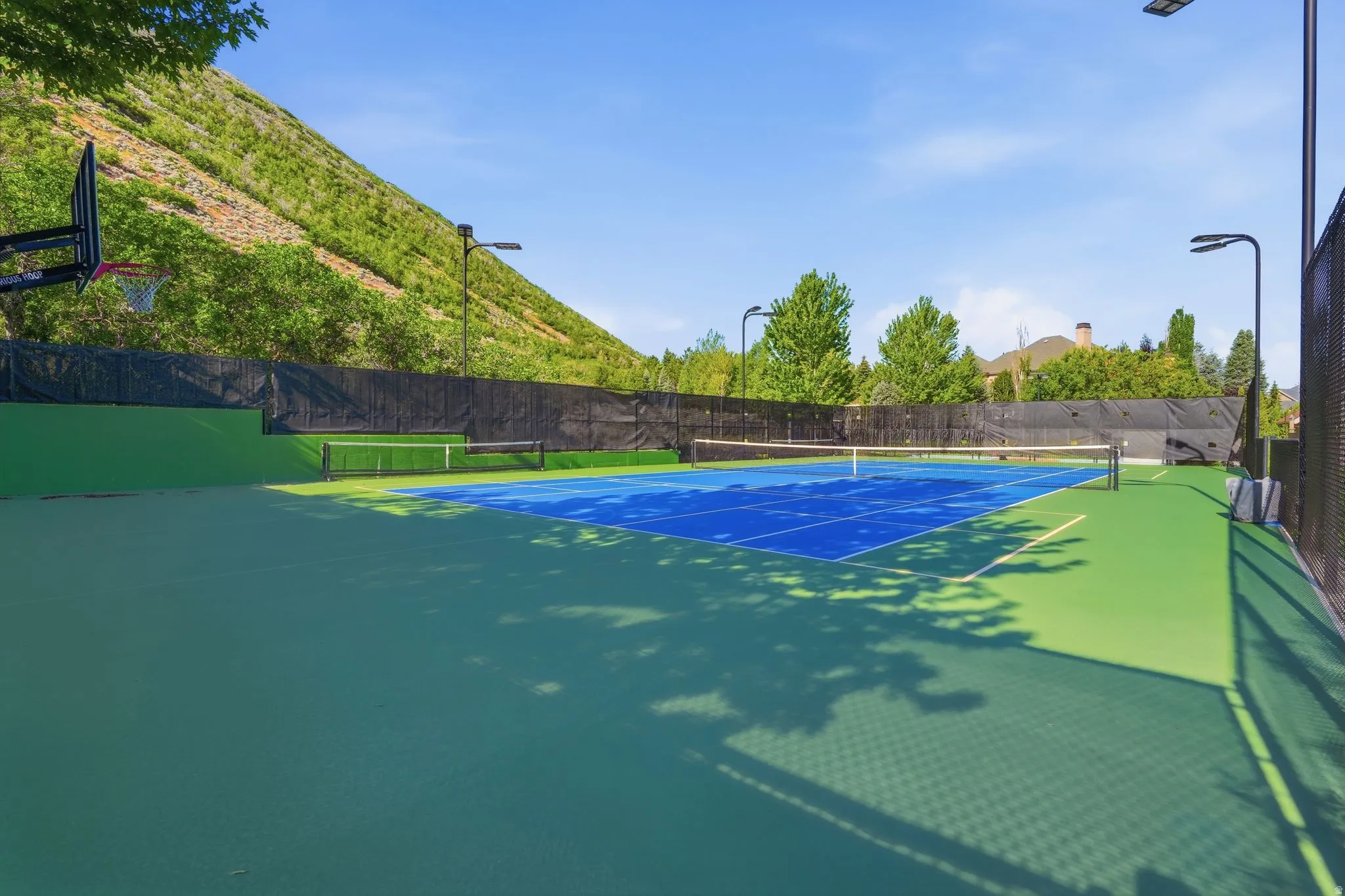 Multi-sport court featuring a blue and green playing surface, Tennis/pickleball, perimeter fencing, overhead lighting, and an integrated basketball hoop