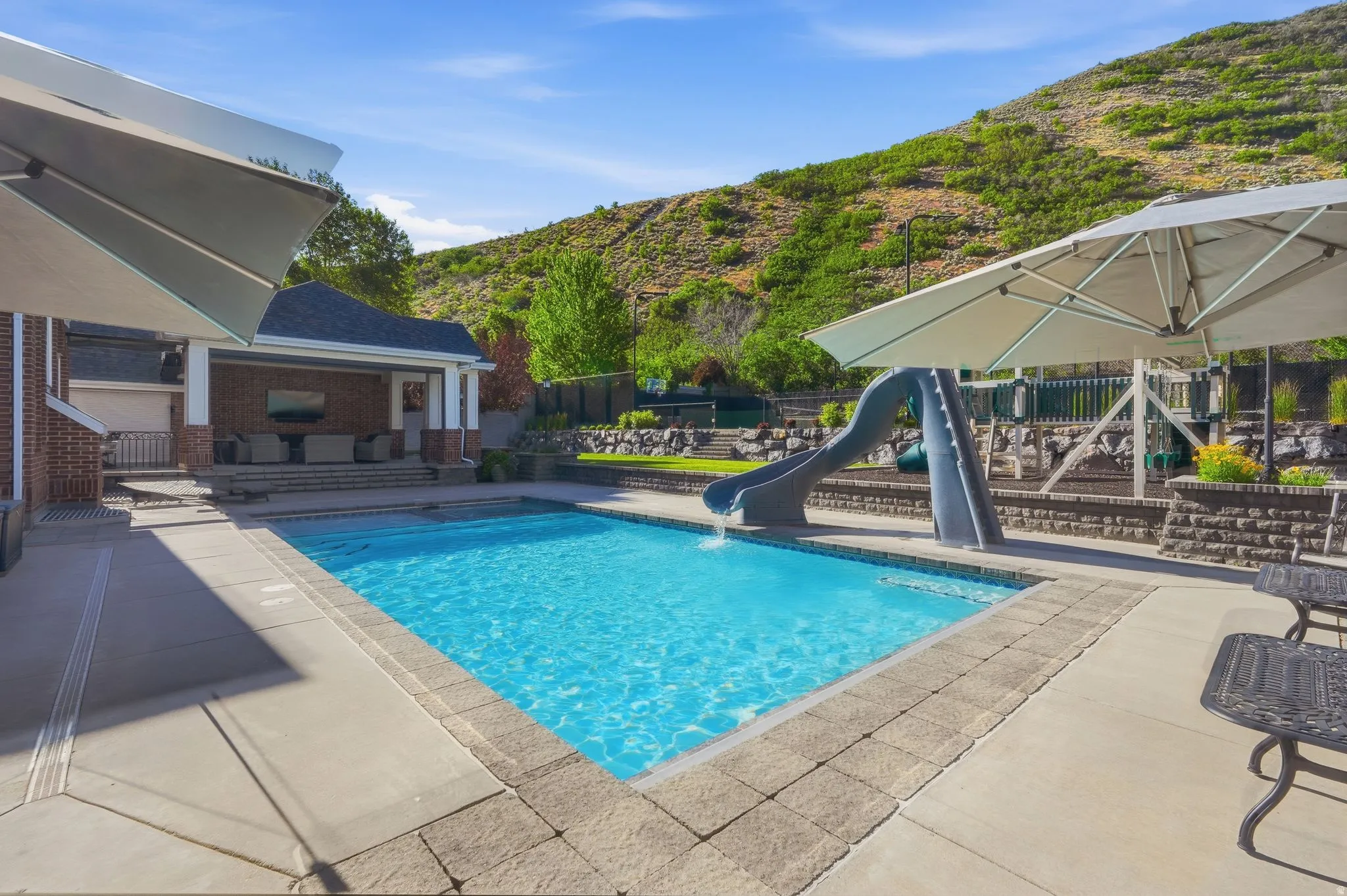 In-ground swimming pool with an integrated hot tup and slide, with included umbrellas and covered patio.