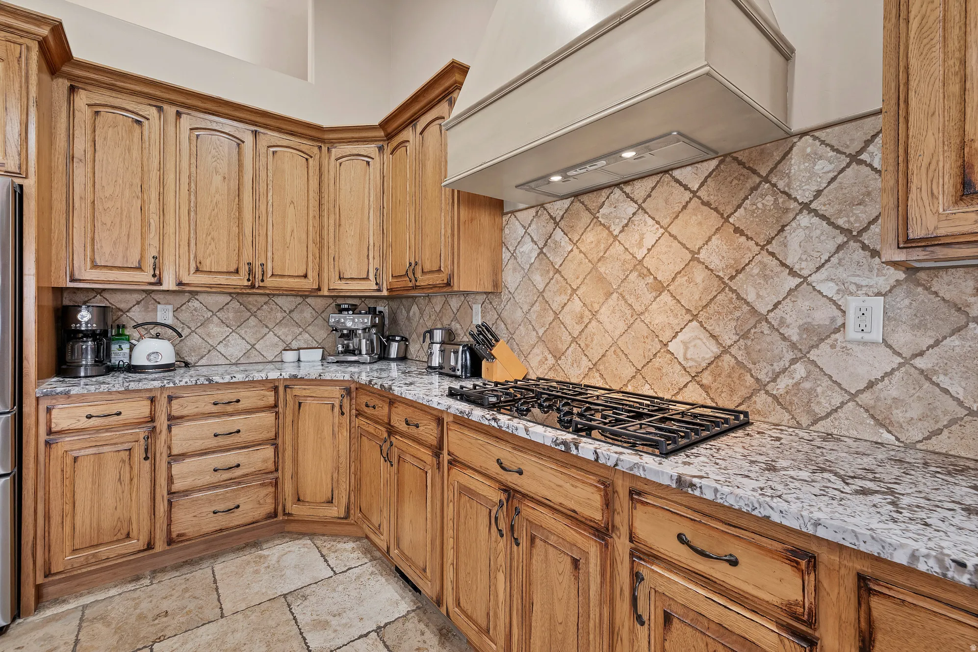 Kitchen featuring light stone countertops, black gas cooktop, wood finish cabinetry, tasteful backsplash, and stone tile floors