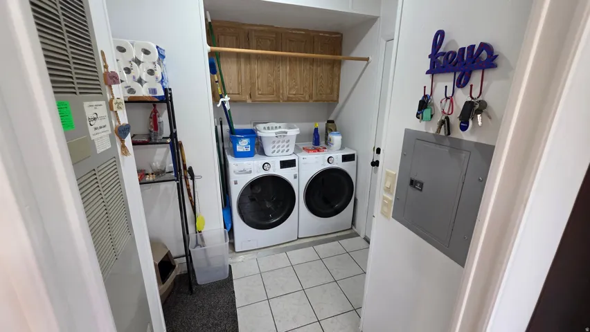 Laundry area with a heating unit, electric panel, washing machine and clothes dryer, light tile patterned floors, and cabinet space
