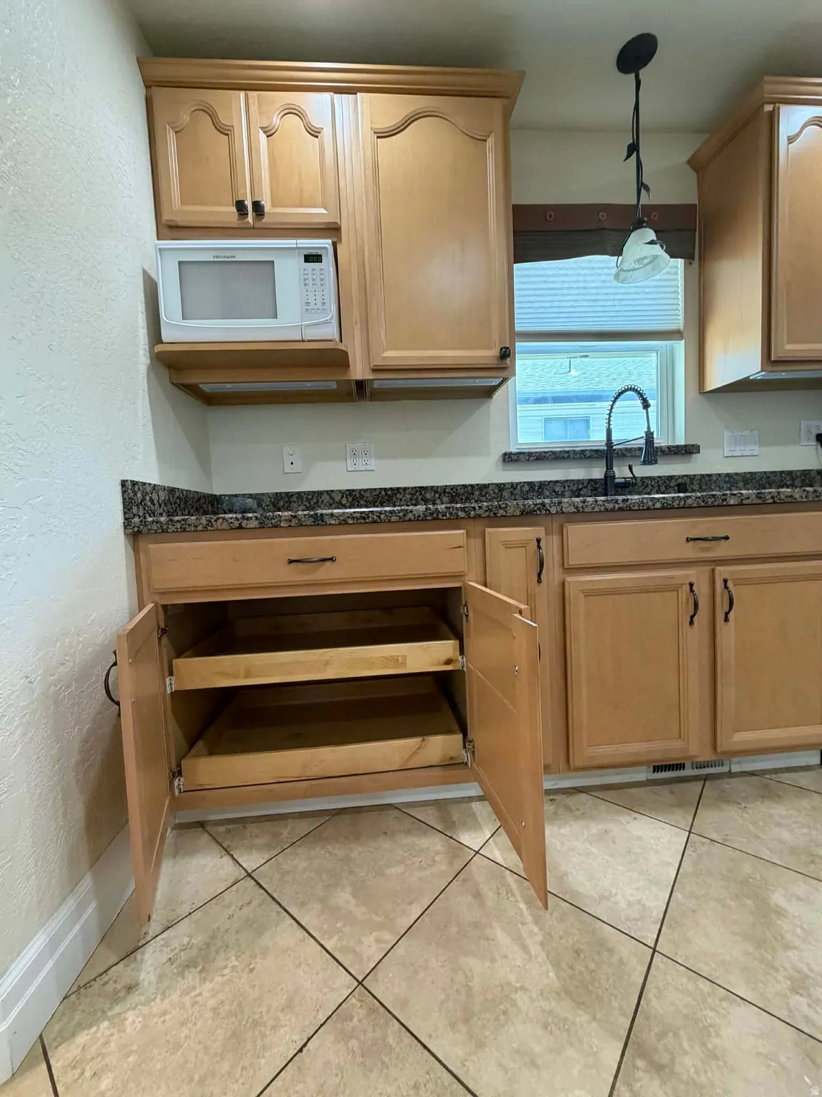 Kitchen featuring a textured wall, white microwave, decorative light fixtures, dark stone counters, and light tile patterned flooring
