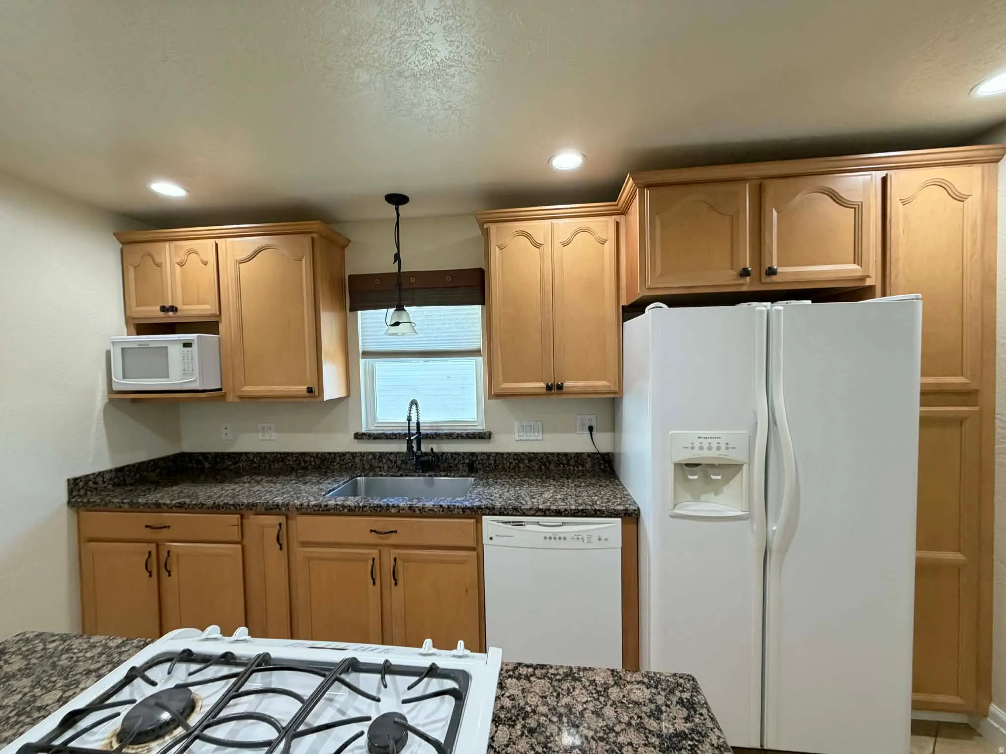 Kitchen featuring white appliances, decorative light fixtures, dark stone counters, and light wood finish cabinetry