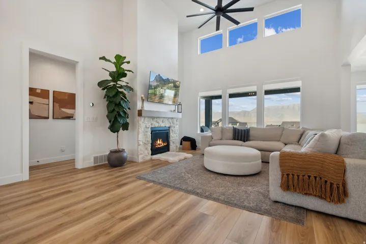 Living room with a stone fireplace, light wood-type flooring, healthy amount of natural light, a high ceiling, and ceiling fan