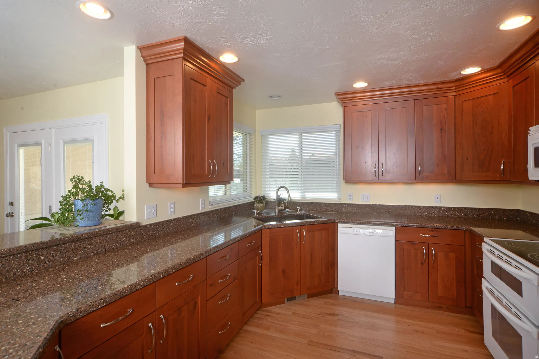 Kitchen with dark stone countertops, white appliances, recessed lighting, wood finish cabinetry, and light wood-style flooring
