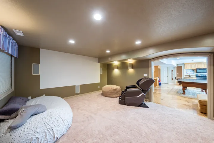 Carpeted recreation room featuring a large projection screen, built-in wall speakers, recessed lighting, and an arched doorway leading to a hardwood-floored area