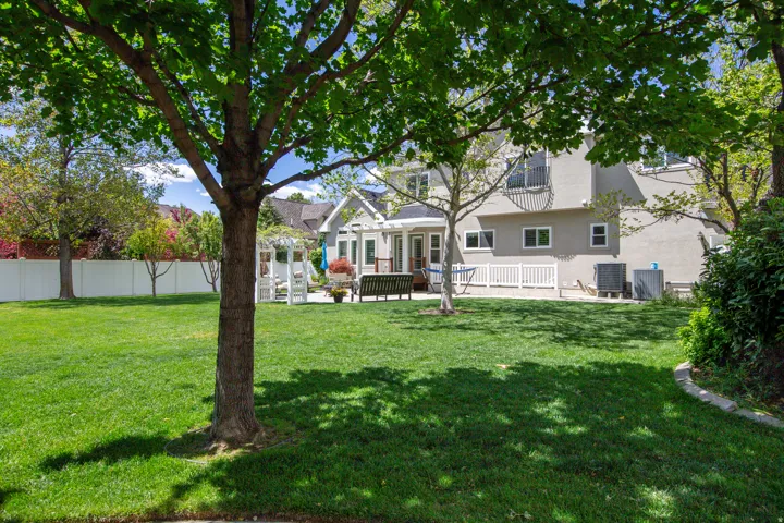 Expansive rear yard featuring a manicured lawn, mature trees, white picket fencing, and a decorative pergola
