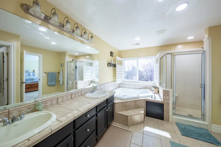 Bathroom featuring a dual-sink vanity with tile countertop, a separate glass-enclosed shower, a corner built-in tub with tile surround, and a vanity light bar with six fixtures
