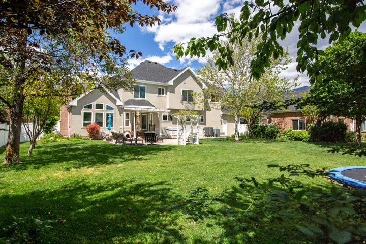 Expansive backyard featuring a large green lawn, mature trees providing natural shade, a stucco exterior with multiple windows, a rear patio, and a white pergola structure