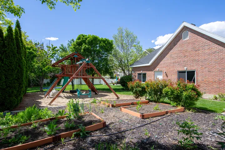 Spacious backyard featuring a wooden play structure with swings and slide, a dedicated sandbox area, multiple raised garden beds, mature trees, and a brick exterior with white trim