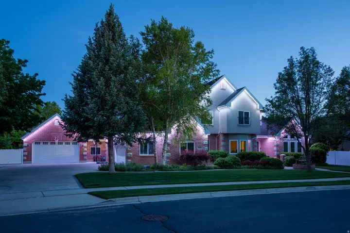 Brick and stucco exterior featuring a two-car garage with white doors, accent lighting, mature landscaping, and a manicured lawn