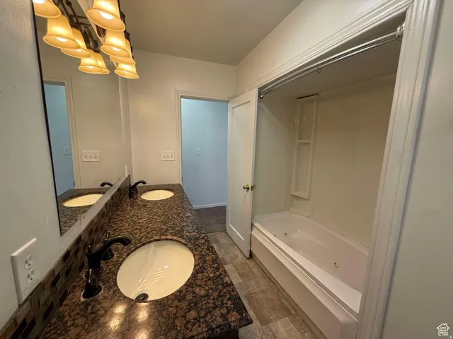 Full bath featuring double vanity, a jetted tub, suspended lighting, and dark stone finish floors