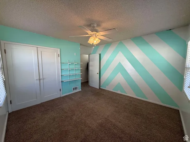Unfurnished bedroom featuring an accent wall, dark carpet, a ceiling fan, wallpapered walls, and a closet