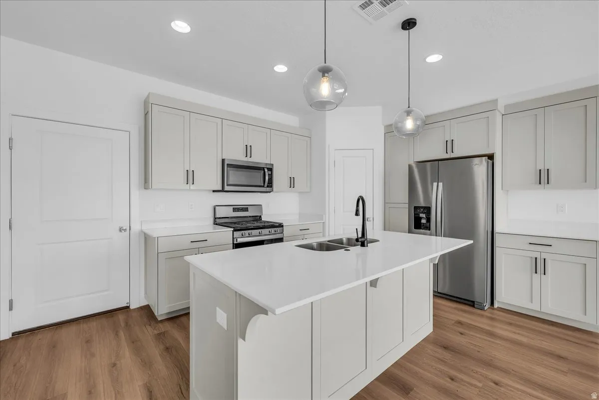 Kitchen featuring stainless steel appliances, decorative light fixtures, light wood-type flooring, a kitchen island with sink, and light stone counters