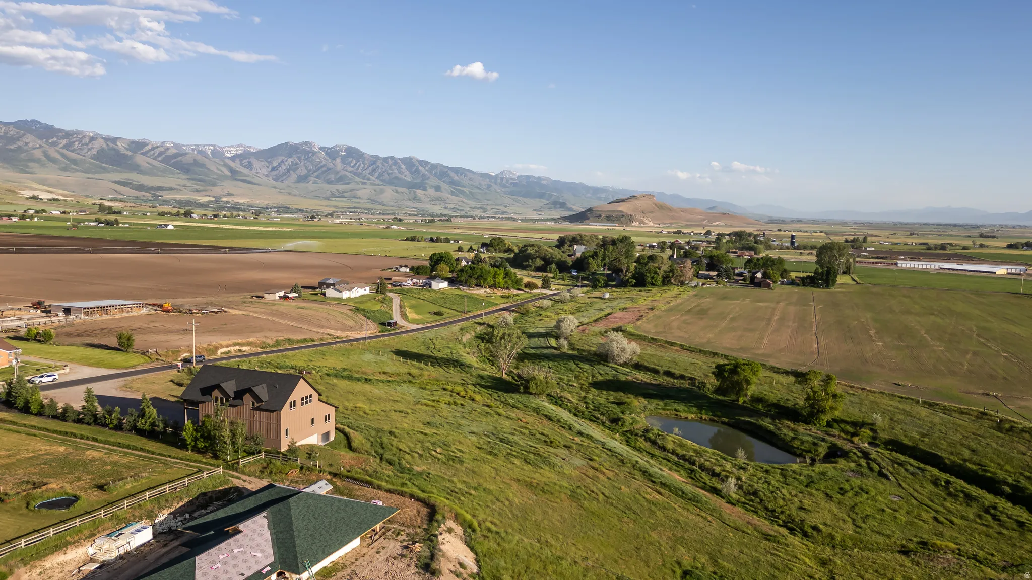 Expansive aerial view showcasing a large residential structure with a dark roof and light-colored siding, surrounded by extensive green and brown fields
