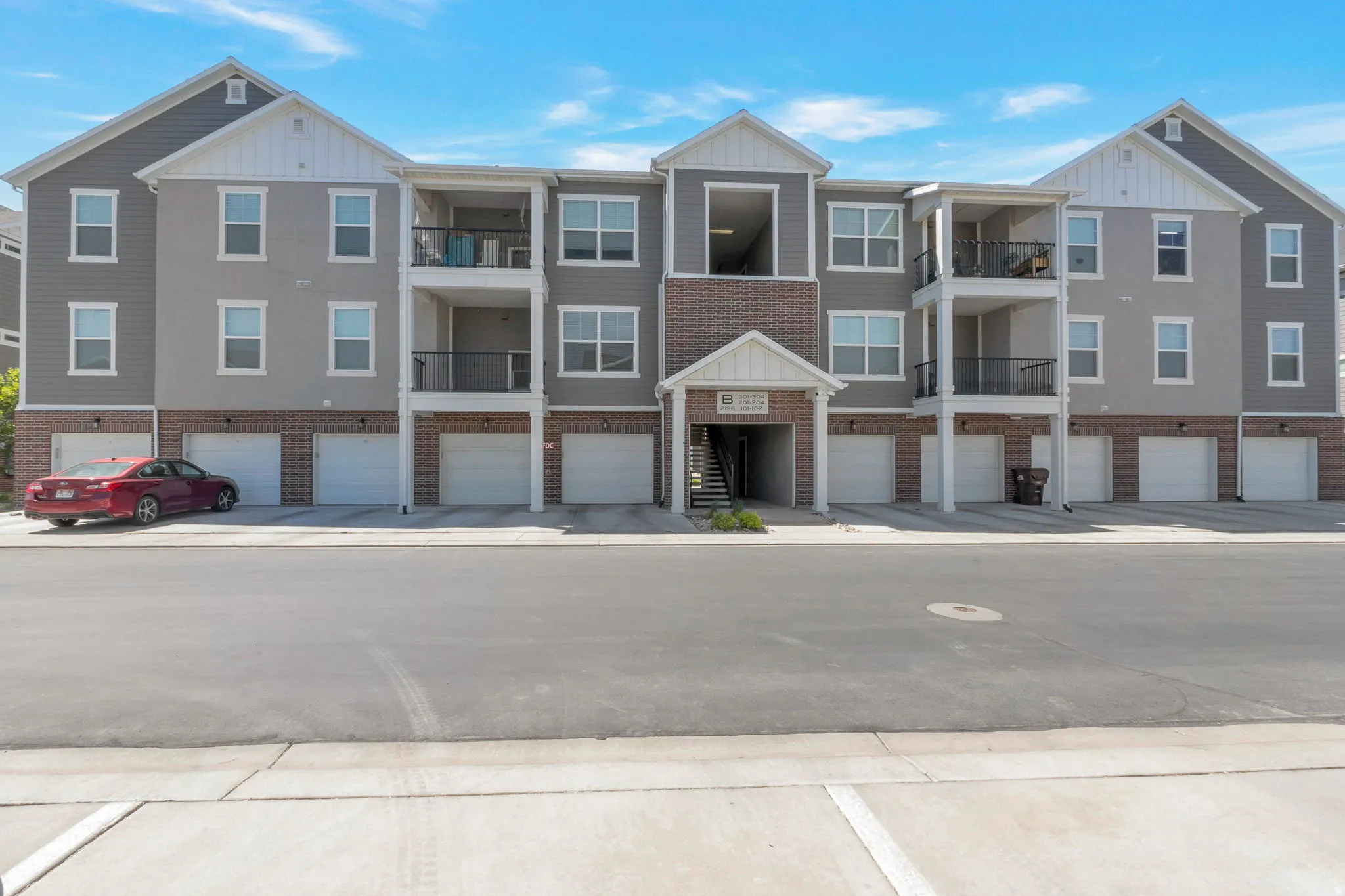 Contemporary multi-story building featuring gray siding, brick accents, white trim, multiple balconies with black railings, and ground-level garages