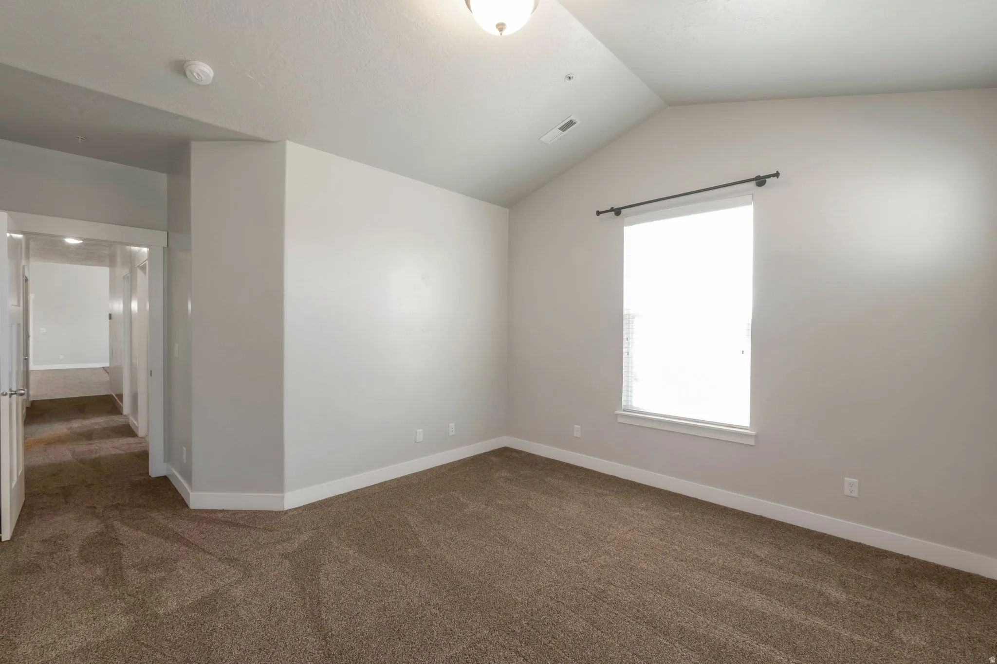 Spacious room featuring vaulted ceilings, a large window with blinds, neutral wall paint, plush carpet flooring, and white trim