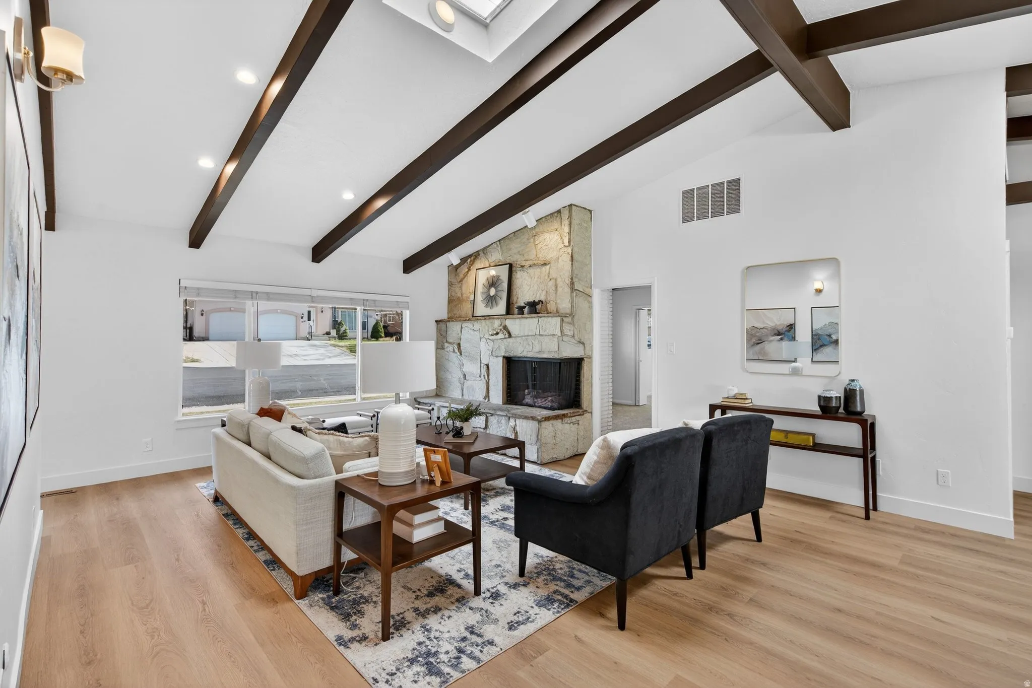 Living area featuring light wood-style floors, vaulted beam ceiling, a stone fireplace, and a skylight