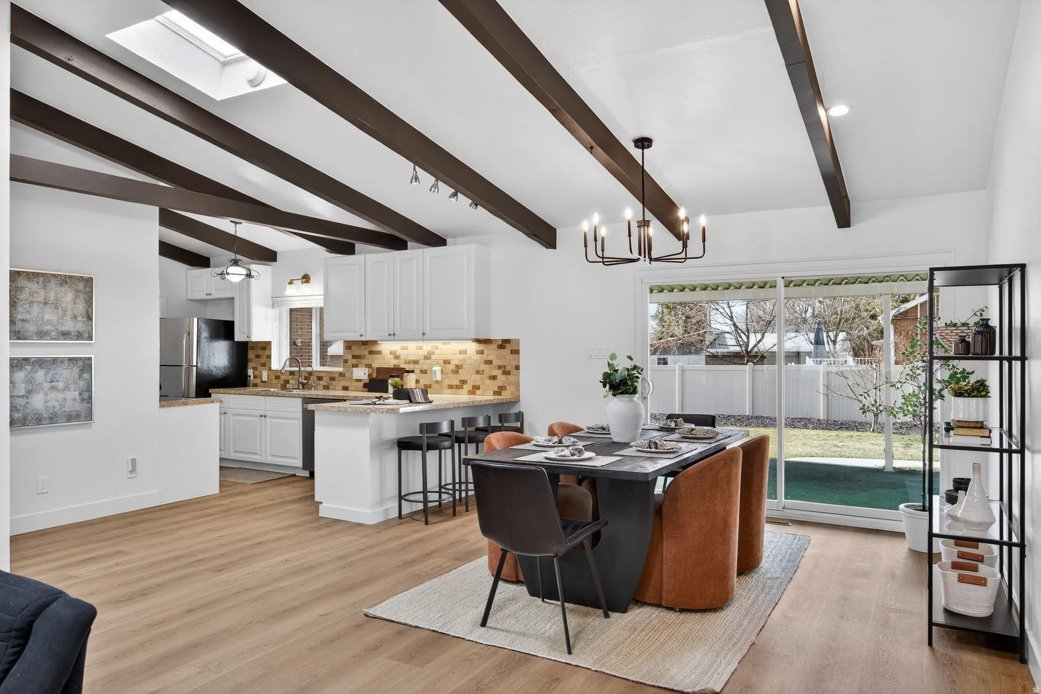 Dining area featuring vaulted beams and a statement chandelier light. Natural light from the skylight bathes the seamless light-toned flooring, highlighting the room's height and open-concept design.