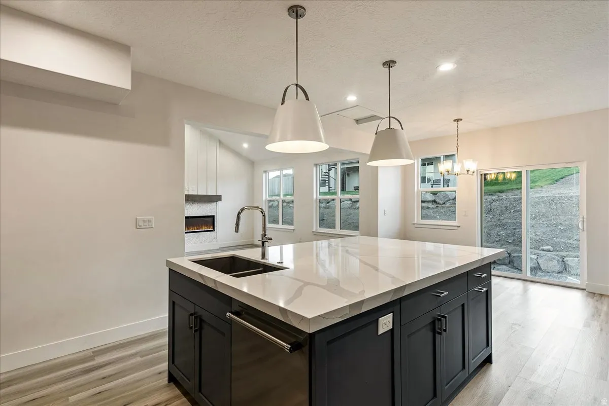 Kitchen with dark cabinetry, dishwasher, a kitchen island with sink, light stone counters, and a large fireplace