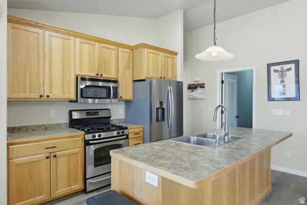 Kitchen featuring light wood finish cabinets, stainless steel appliances, a center island with sink, vaulted ceiling, and hanging light fixtures