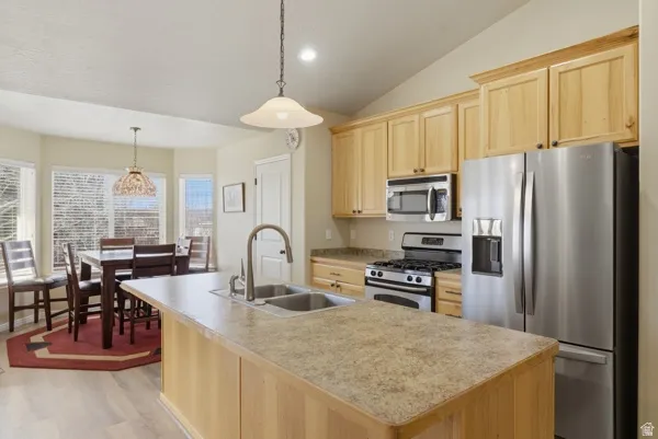 Kitchen with light wood finish cabinets, stainless steel appliances, hanging light fixtures, a kitchen island with sink, and light countertops