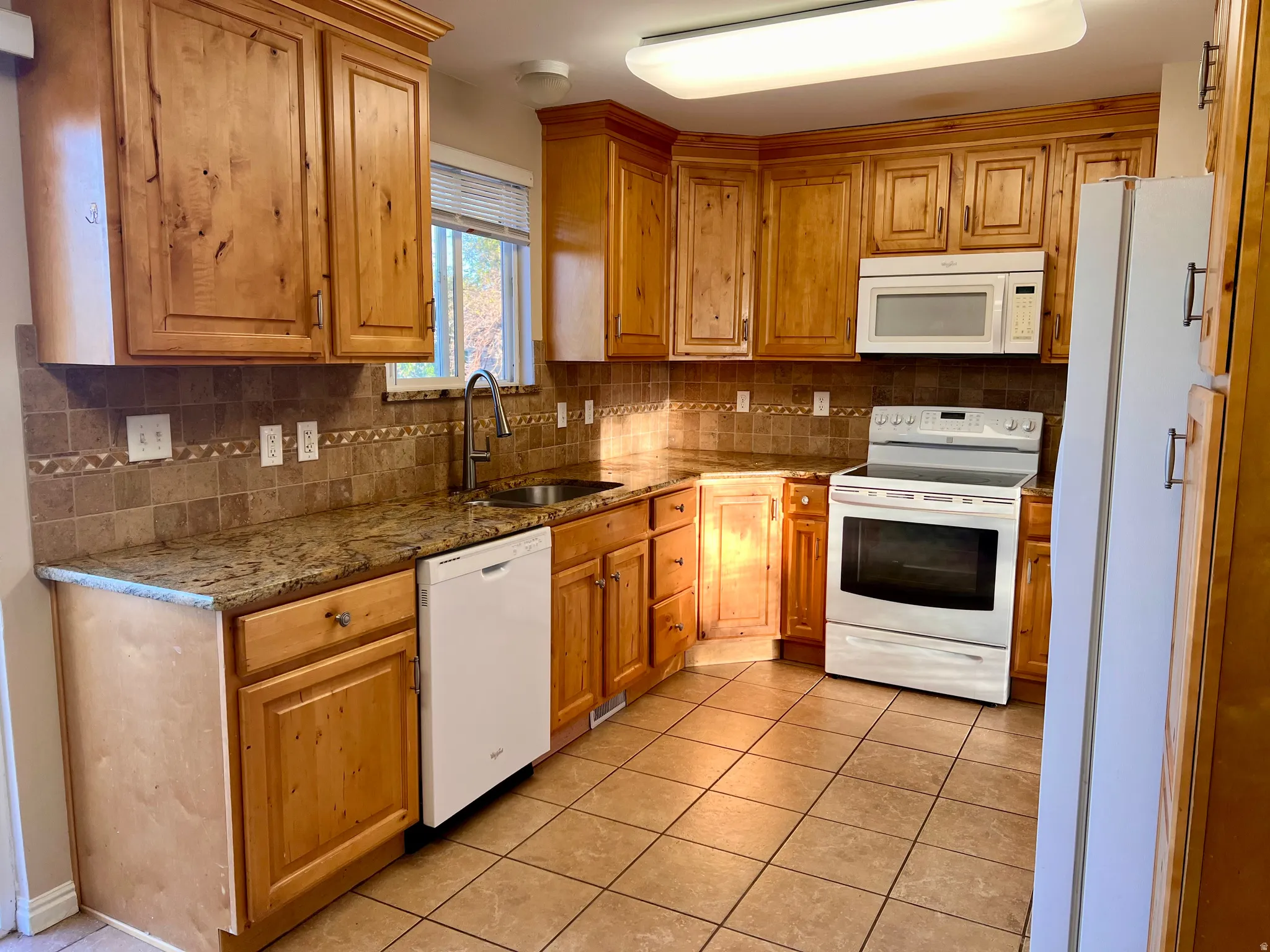 Kitchen with white appliances, dark stone counters, wood finish cabinetry, light tile patterned floors, and decorative backsplash