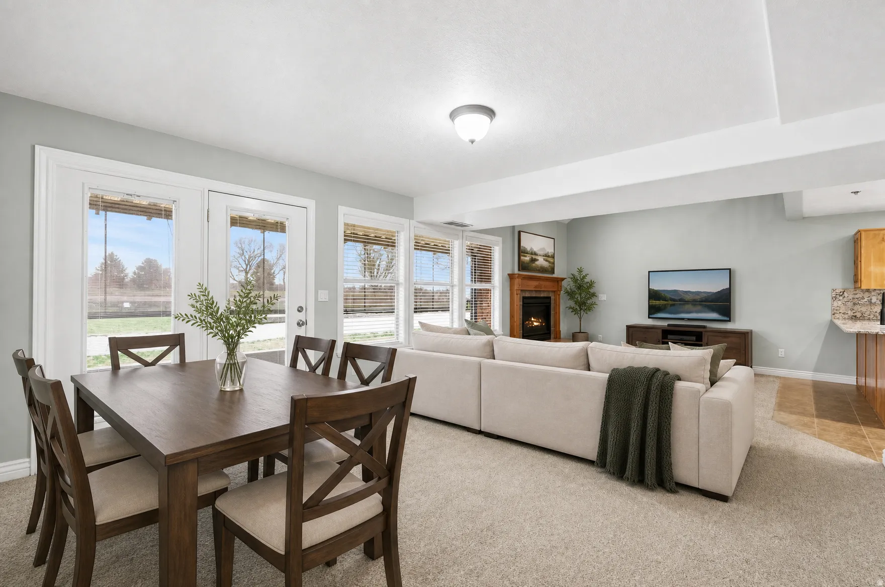 Open-concept living area featuring a central fireplace with wood mantel, light-toned carpeting, and a dining space with direct outdoor access