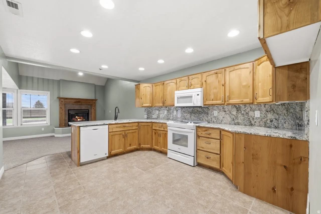 Kitchen featuring a peninsula, backsplash, white appliances, a warm lit fireplace, and recessed lighting