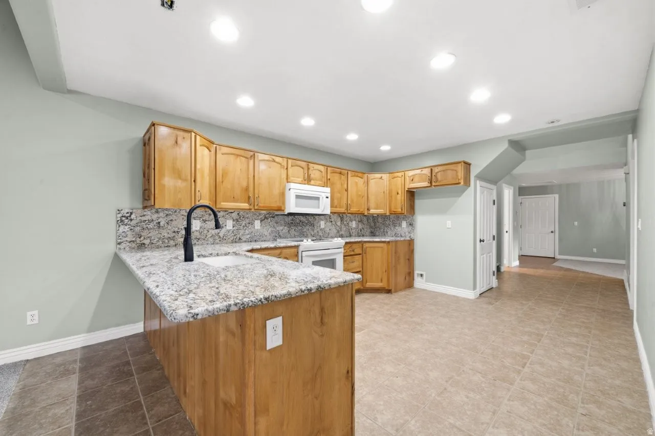 Kitchen with backsplash, a peninsula, light stone counters, white appliances, and recessed lighting