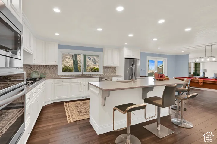 Kitchen with stainless steel appliances, a kitchen bar, white cabinets, an island with sink, and dark wood-type flooring