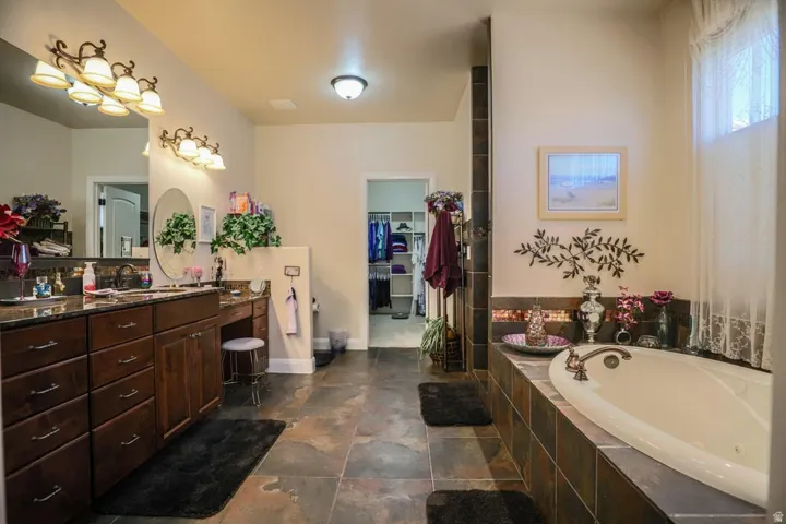 Dual vanity with granite countertop and dark wood cabinetry, large soaking tub with tile surround, walk-in closet, and tile flooring