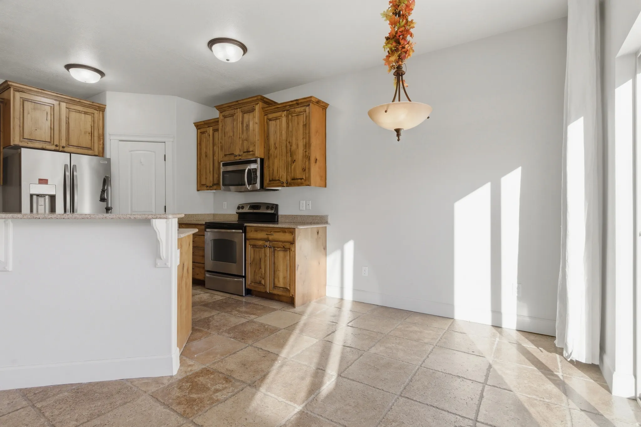 Kitchen featuring stainless steel appliances, stone tile flooring, wood finish cabinets, pendant lighting, and light stone counters