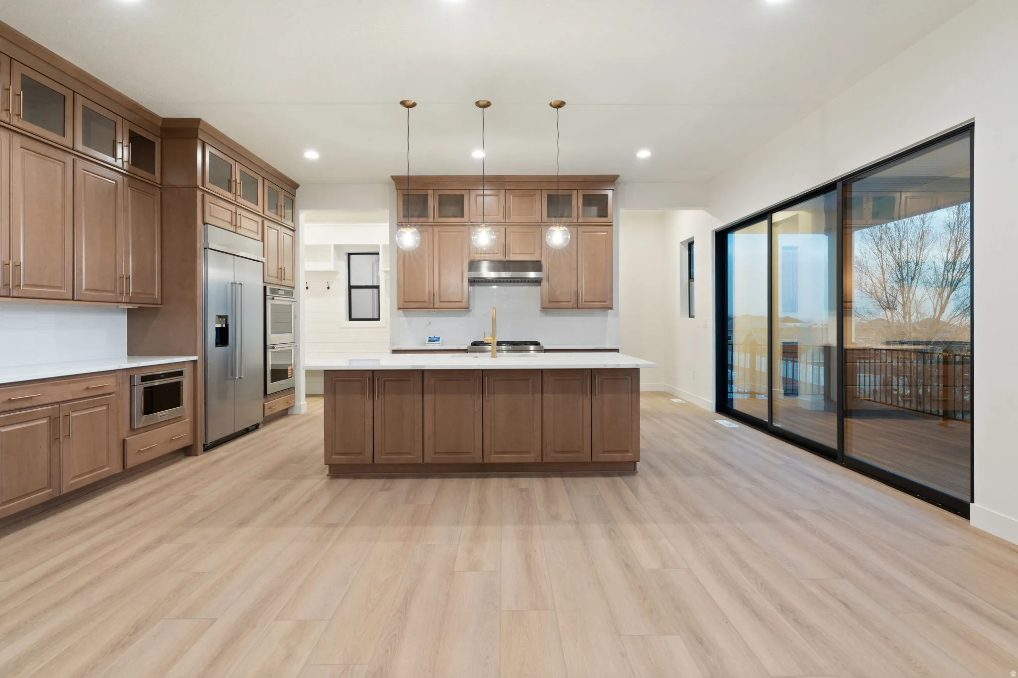 Kitchen featuring stainless steel appliances, a kitchen island with sink, wood finish cabinets, hanging light fixtures, and light wood-type flooring