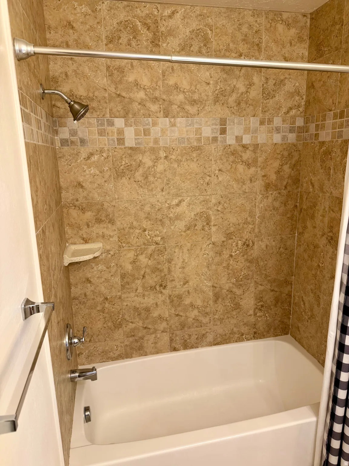 Bathtub and shower combination featuring neutral-toned wall tiling, decorative mosaic accent band, and chrome fixtures